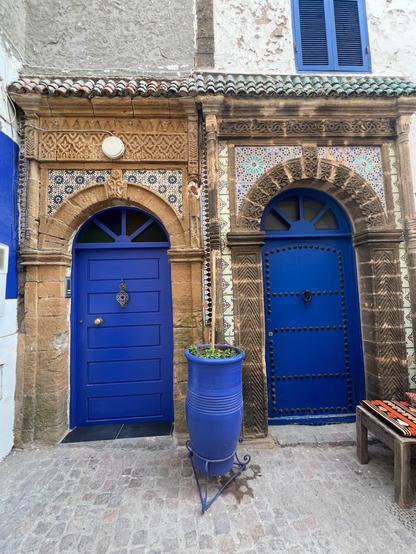 Two vibrant blue Moroccan doors are side-by-side in an aged stone building. Both are set in elaborately carved stone archways, adorned with intricate blue and white mosaic tiles. The door on the left has a golden knocker, while the door on the right is studded with dark circles. A tall, fluted blue pot stands between the doors.