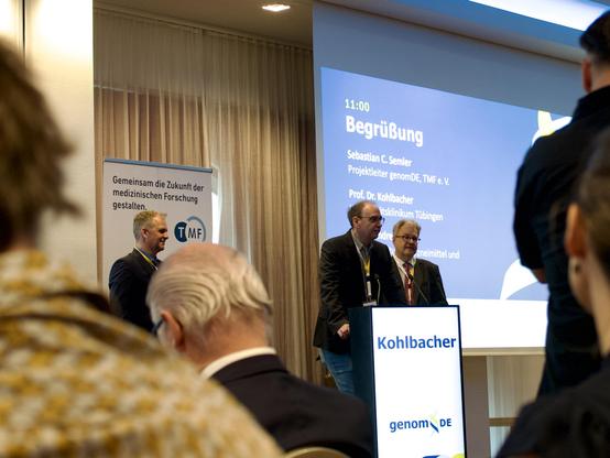 Picture of the welcome address at the genomDE symposium, showing Oliver Kohlbacher speaking with Andreas Till and Sebastian Semmler in the back.
