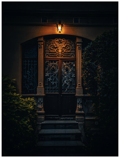 A photo of a doorway at late evening. The doorway is ornately framed with stone and metal detailing, and features a partially visible wooden door with intricate wrought iron metalwork. There isa  small, lit lantern which hangs on the wall above the door.  The steps leading to the door are visible, and lush green plants frame the doorway on either side.