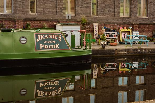 EN: A striking green houseboat (named Jeanie's Pride) is moored in the Union Canal in Edinburgh. In the background you can see the stone row of houses on which colourful objects have been artistically draped (e.g. a light blue bench, a string of lights and some flower pots). All of this is reflected on the smooth surface of the water.

DE: Ein auffällig grünes Hausboot (namens Jeanie's Pride) liegt im Union Canal in Edinburgh vor Anker. Im Hintergrund sieht man die steinerne Häuserzeile, an welcher bunte Gegenstände künstlerisch drappiert wurden (z.B. eine hellblaue Sitzbank, eine Lichterkette und einige Blumenkübel). Das alles spiegelt sich auf der glatten Wasseroberfläche.