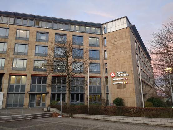 Sechsstöckigen Bürohaus mit großen Fenstern. Dazu an einer Fassadenseite ein weißes großes A (Zeichen der Agentur für Arbeit), dazu auch der Schriftzug "Agentur für Arbeit" sowie "Jobcenter"