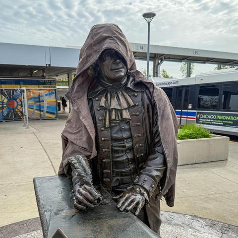 A bronze statue of a historical figure in a hooded cloak, positioned next to a table with a quill. The background features a bus stop and colorful signage.