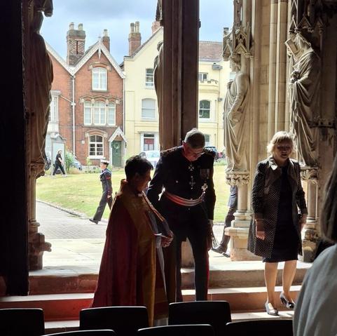 A uniformed man speaks with a robed vicar at the entrance to a cathedral. They are backlit by the open doors to the outside.