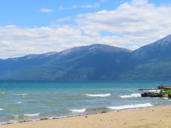 View of the sandy lakeshore, wind-whipped lake waves, and the mountains on the Albania - North Macedonia border, which are only wooded at the bottom. The tops are rounded, eroded stone and appear to be deep blue and purple and blue-green. The lake's waters are sea green, blue-green, and blue.