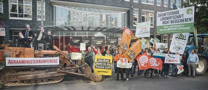 Links im Foto sieht man eine orange Planierraupe mit der Aufshrift "Agrarministerkonferenz" und als MInister verkleidete Personen darauf.Rechts Aktivist*innen, die sich dieser in den Weg stellen. Transparente verschiedener Organisationen sind zu sehen, u.a. AbL, SlowFood, NaJu, Greenpeace, Bioland und ProVieh.