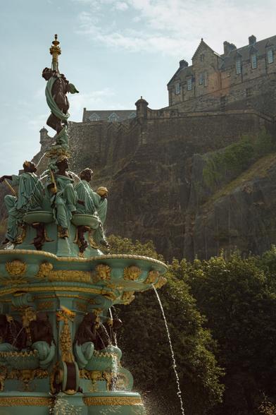 EN: On the left in the foreground, the gold-decorated turquoise-green fountain from the 19th century bubbles away. It consists of several figures and basins, from which the water splashes down again over several storeys. The picture is framed so that the castle walls of Edinburgh Castle (in the background on the right) are (halfway) flush with the fountain.

DE: Links im Vordergrund sprudelt der goldverzierte türkis-grüne Brunnen des 19. Jahrhunderts. Er besteht aus mehreren Figuren und Becken, aus denen das Wasser über mehrere Stockwerke wieder hinunterplätschert. Der Bildausschnitt ist so gewählt, dass die Burgmauern des Edinburgh Castle (rechts im Hintergrund) mit dem Brunnen (halbwegs) bündig einen Abschluss finden.