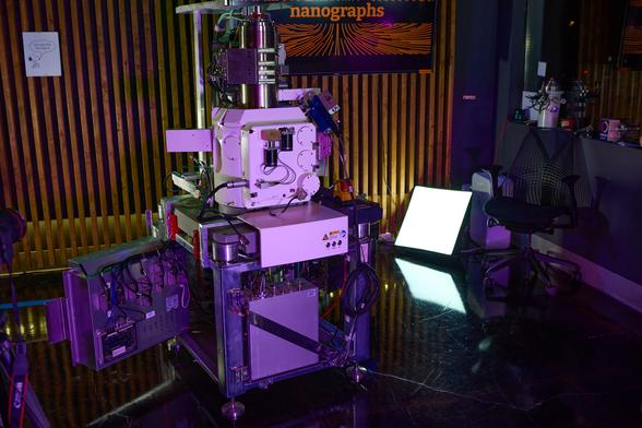 Picture of Zeiss Merlin electron microscope from the front with a TV in the background with the Nanograph logo on it.