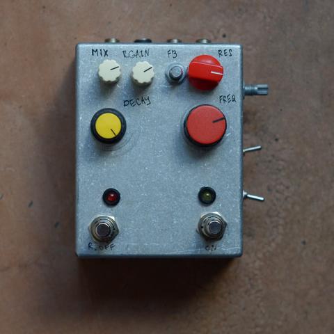 A photo of an effect pedal. All the knobs are different sizes and colors, a bunch of switches on the right side of the box. Knobs are signed by hand: Mix, R.Gain, FB, Res, Decay, Freq. Two soft footswitches.