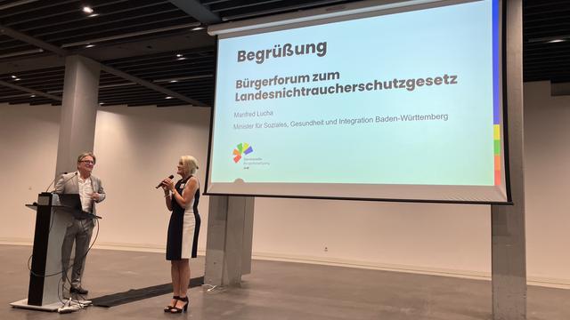 Minister Manne Lucha und eine Frau stehen vor einer Leinwand. Vor Minister Manne Lucha steht ein Rednerpult. Die Frau spricht in ein Mikrofon. Im Hintergrund sieht man eine Folie mit der Aufschrift "Begrüßung. Bürgerforum zum Landesnichtraucherschutzgesetz".