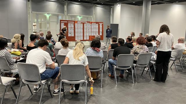 Man sieht viele Menschen, die auf Stühlen sitzen. Vorne stehen eine Frau und ein Mann vor einer roten Pinnwand. Die Frau hat ein Mikrofon in der Hand. Auf der Pinnwand sind viele weiße Zettel angepinnt.