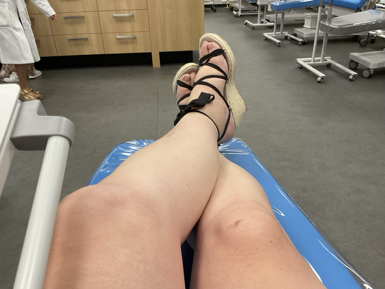 Emma zit ontspannen met haar benen gekruist tijdens het doneren van plasma bij de bloedbank. Ze draagt espadrilles met een hak met zwarte linten.