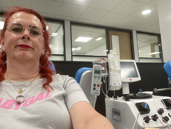 Emma, met rood haar en bril, glimlacht ontspannen tijdens haar plasmadonatie bij de bloedbank. Achter haar zijn medische apparaten te zien die gebruikt worden voor het afnemen van plasma. Ze draagt een grijs T-shirt met in roze letters "BABYGAL"