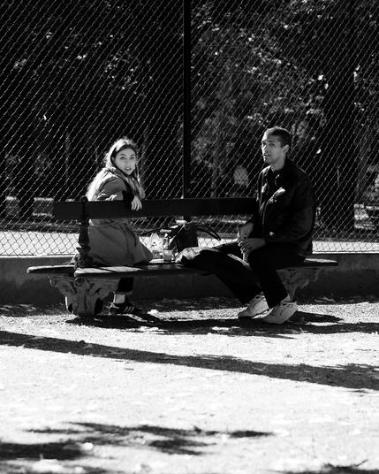 Two people sit on a park bench beside a chain-link fence. The person on the left wears a coat and looks towards the camera, while the person on the right is dressed in a dark jacket and pants. A bag and a bottle are on the bench between them. The ground is covered with gravel and dappled with shadows. The image is in black and white.
