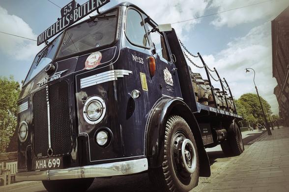 A low-angle, wide shot of a vintage dark blue Mitchells & Butlers delivery truck, number 53, with wooden barrels loaded in the back. The truck has a distinctive large grille and round headlights, with "MITCHELLS & BUTLERS" prominently displayed above the windshield. It is parked on a cobbled street with a building and trees visible in the background under a cloudy sky. The image has a slightly desaturated, vintage feel.