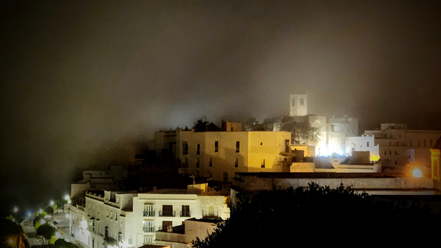 Wolken ziehen durch einen Ort auf einem Hügel. Die weißen Häuser liegen im Nebel, der in der Dunkelheit von den Lichtern illuminiert werden.