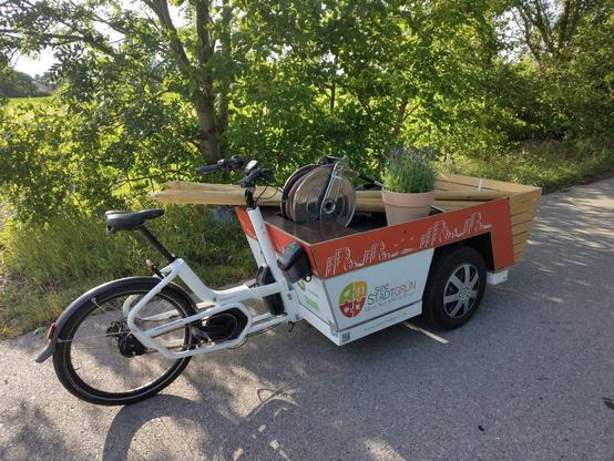 Das Gießrad von Seestadtgrün steht am Rande von einem asphaltierten Weg. Beladen ist es mit mehreren Holzpfählen und einem Topf mit Lavendel. Die Sonne scheint durch die Bäume am Wegesrand und erzeugt so einen sommerlichen Schatten am Weg.
