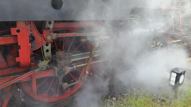 Das Bild zeigt den unteren Teil einer Dampflokomotive. Es ist viel Dampf zu sehen, der die rot-schwarzen Bauteile teilweise verdeckt.
