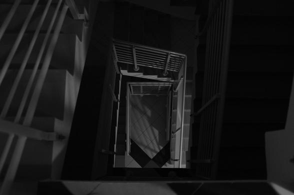 Looking down a narrow, rectangular spiral staircase in black and white, with a repetitive pattern of railings and steps creating geometric shapes. The floor at the bottom has a tiled pattern, visible through the center of the spiral. Shadows and lighting accentuate the stairway’s railing and step outlines.