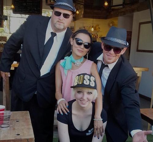 Medium shot of four people posing formally for a camera. Two men, positioned to the left and right of the frame, wear dark suits, white shirts, black ties, hats, and sunglasses. In the center between them a woman standing and wearing sunglasses, a turquoise-blue and rhinestone necklace, a pink top, & earrings; a smiling blond-haired woman sits in front wearing a "BOSS" hat. The background is a blur of a cafe-style indoor setting with soft daylight