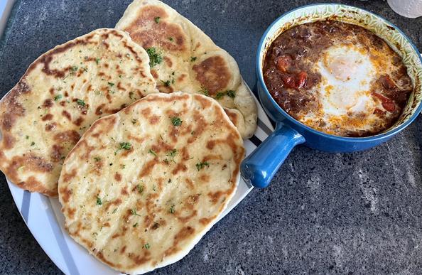 a plate of three flatbreads in the style of garlic naan, with lots of crispy bits and a scattering of herbs. to one side is a heavy oven-proof bowl of dark meaty stew with two eggs that have been poached in the sauce