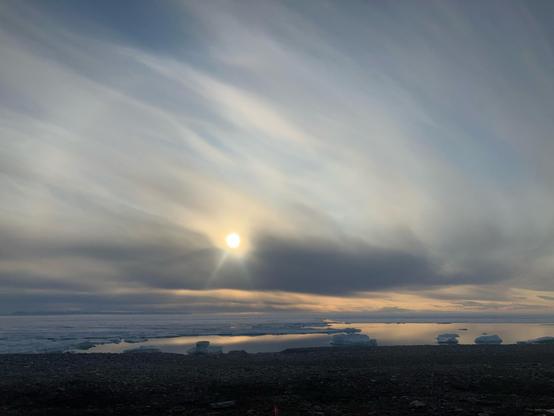 A shoreline of the Arctic Ocean. A open area of dead calm water, framed by sea ice, reflects orange from the sun lit sky. The sun is low and partially obscured by clouds. On either side of the sun are faint sun dogs. And joining them in an arc is an arc of light