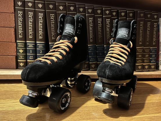 A pair of black suede high top quad roller skates, black wheels, and orange laces sit on a wooden floor in front of a bookcase holding volumes of the Encyclopedia Brittanica.