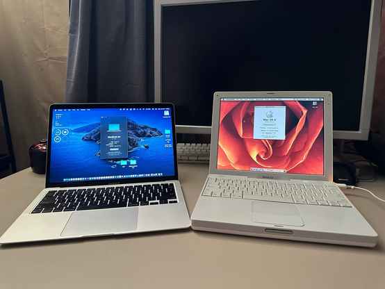 Two Apple laptops are side by side on a beige desk: on the left is a more modern M1 MacBook Air in silver, and on the right is a white iBook G4 running Mac OS X Tiger. The MacBook Air displays the macOS Sequoia desktop, with the “About This Mac” window open, showing it as a MacBook Air with an Apple M1 chip and 8GB of RAM. The iBook G4 screen also shows its system information, revealing it’s running Mac OS X 10.4.11 with a 1.2GHz PowerPC G4 processor and 1.25GB of RAM. The iBook has a vivid rose-themed wallpaper and is plugged into power, with its charging indicator glowing orange.  Behind the laptops is a large 24-inch Sun Microsystems LCD monitor, currently powered off, and a white Apple keyboard tucked partially underneath it. A Microsoft-branded mouse is visible on the right edge of the desk, and a partially obscured camera sits to the left of the MacBook Air.