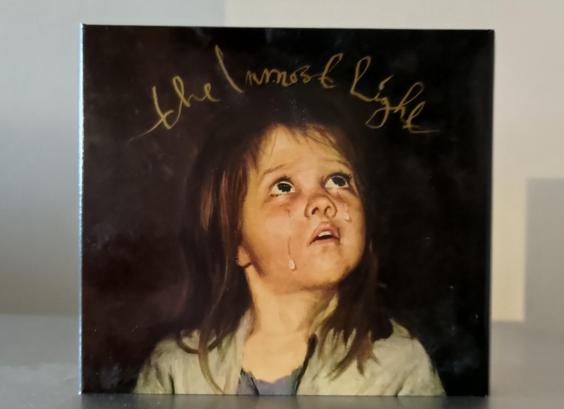 Digipak CD cover with a painting of a girl crying, tear on her cheek, looking upwards. Dark brown hair, light skin. Dark brown background. 

"The Inmost Light" handwritten in gold over her head.