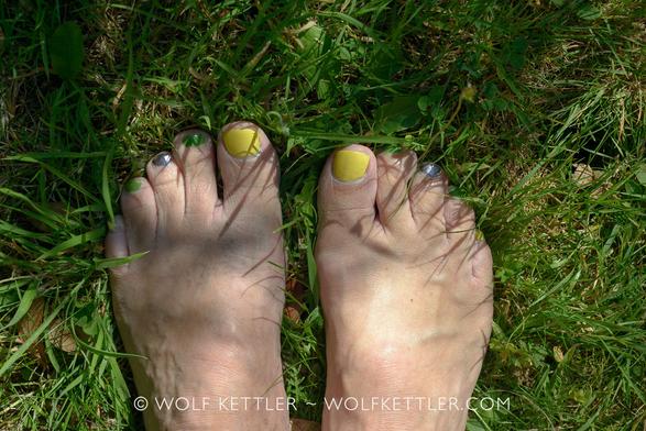 Photo of a man’s bare feet with painted toenails, standing in the grass. The colour scheme is yellow, green and metallic purple.