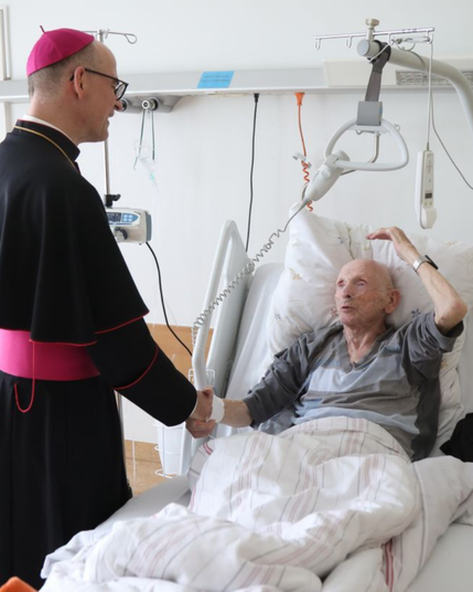 Ein Mann liegt in einem Krankenbett, der Bischof hält seine Hand.