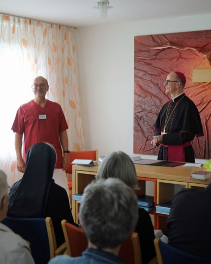 Der Bischof hinter einem Tisch, neben ihm steht ein Pfleger und spricht, vor ihm sitzen einige Leute auf Stühlen.
