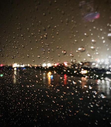 Photograph of airport yellow, blue and red lights seen through a plane window, on a rainy night. The focus is on the raindrops on the surface of the window.