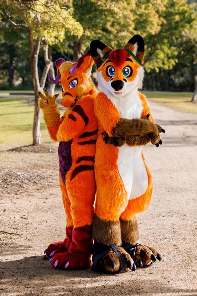 Lino Plum (orange, purple and maroon dragon fursuiter) in full fursuit standing with one arm folded and the other showing the number 3, while back-to-back with Colto Fox (taller orange, white, brown and red fox fursuiter) also in full fursuit standing with his arms crossed wearing a green beanie.