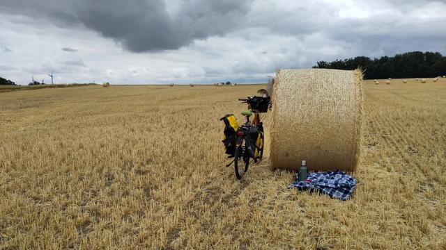 Fahrrad auf einem Stoppelfeld, angelehnt an einem Strohballen. Davor eine Jacke auf dem Boden , eine Thermoskanne und eine rote Blechtasse. Der Himmel ist bewölkt