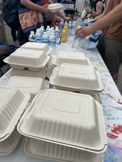 A stack of closed food containers on the distro table. Beyond the trays you can see water bottles and people giving and accepting accepting the food and water.