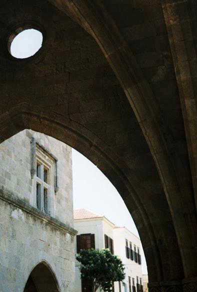Color photograph looking out from under a medieval style stone arch. A small circular window is above the point of the arch. Stone buildings are visible in the background.