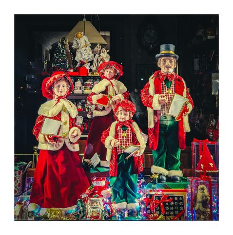 This photo shows a Christmas window display featuring four life-sized figurines: a man in a top hat and green pants, two girls in red dresses, and a boy in a red and green plaid outfit. All four figures hold songbooks.  The display is set against a backdrop of a Christmas tree, other decorations, and wrapped gifts. The figures are dressed in traditional Christmas clothes. A small Santa figurine and a calendar are visible in the foreground. The figures' expressions are serene and they seem to be a family of carolers. Underneath them on the display there is an advent calendar, a Santa Claus and other festive items made out of chocolate.