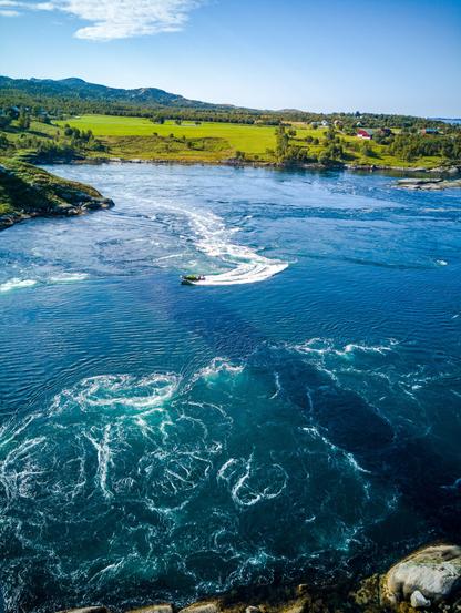 Photo shows a narrow fiord with visible strong vortices and whirlpools, and small boats crisscrossing in and out of them. The photo is taken from above, from the road bridge crossing this current. The water is deep blue-green, the landscape is variations of green with some start of fall red orange colours. In the background low mountains, rolling hills and a small farm. To the right there are glimpse of the larger fiord feeding the Maelstrom with water.