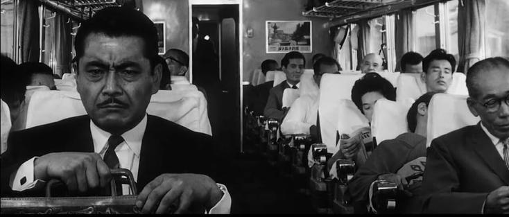 Black and white photo still from the 1963 Kurosawa film High and Low. An intenese looking man with a moustache staring forward on the left of the frame, sitting in a very busy train.