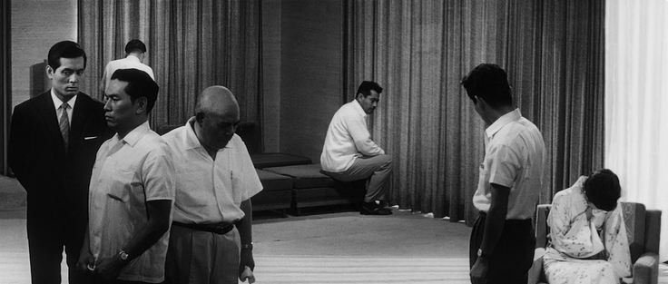 Black and white photo still from the 1963 Kurosawa film High and Low. One woman in a kimono sitting down on the lower right, looking very upset as well as six men spread across a minimalist mid-modern living room.