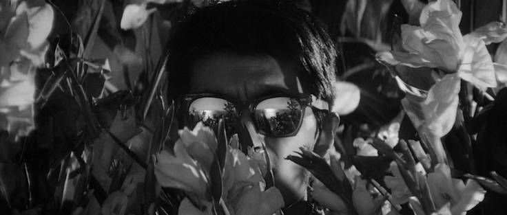Black and white photo still from the 1963 Kurosawa film High and Low. Facing the camera, a man with reflective Ray Bans is peering through a bunch of flowers