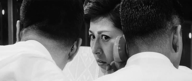 Black and white photo still from the 1963 Kurosawa film High and Low. A woman with a concerned expression is looking toward the camera. She is seen between the backs of two men's heads with very short hair and white shirts. The man on the right is holding a phone receiver to his ear.
