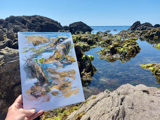 Hand holding watercolour sketch in front of rockpools and sea with blue sky