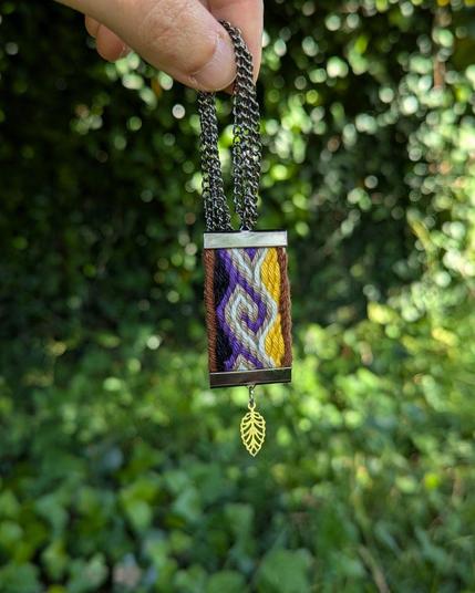 Photo d'un pendentif tissé aux couleurs du drapeau non-binaire tenu à deux doigts devant un mur de lierre.