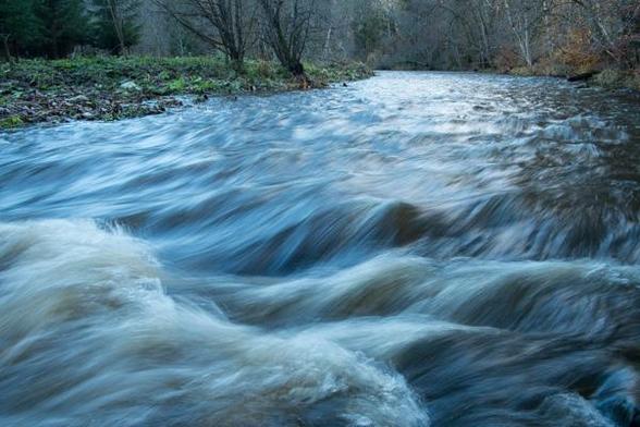Ein Fluss mit einer starken Strömung und Wellengang wurde aus einem niedrigen Winkel knapp über der Wasseroberfläche fotografiert, mit Blick stromaufwärts gegen die Strömung. Aufgrund der leicht verlängerten Belichtungszeit erscheinen die Wellen weichgezeichnet, und das Wasser zeigt streifenartige Muster, die die Dynamik des Flusses betonen. Man hat den Eindruck, das Wasser ströme dem Beobachter direkt entgegen.