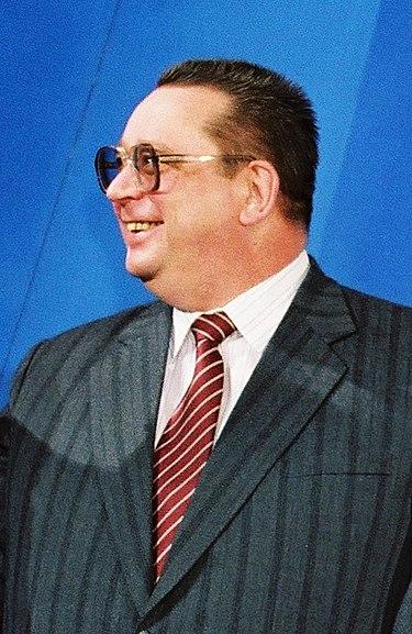 50ish yr old man. Light skin tone. Dark hair. Wearing a striped suit and a red tie. Has glasses and a large ear.
