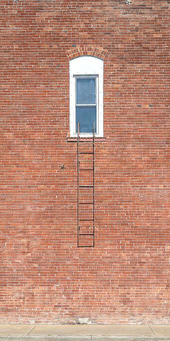 A single second story window in brick building has a short metal fire escape ladder hanging straight down below it to about 5 or 6 feet off a sidewalk.