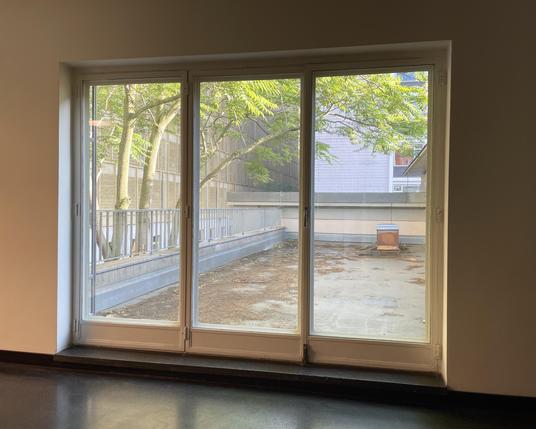 View from inside a building through three large windows or maybe even doors. The terrace outside is empty except for two bee hives in the back. The terrace is on the first floor of the building so next to it there are tree trunks and the lower branches visible.