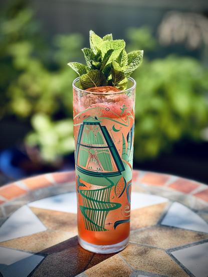 a bright red cocktail with long clear ice spear and mint garnish in a B-Rex zombie glass sitting on a table in partly shady sun