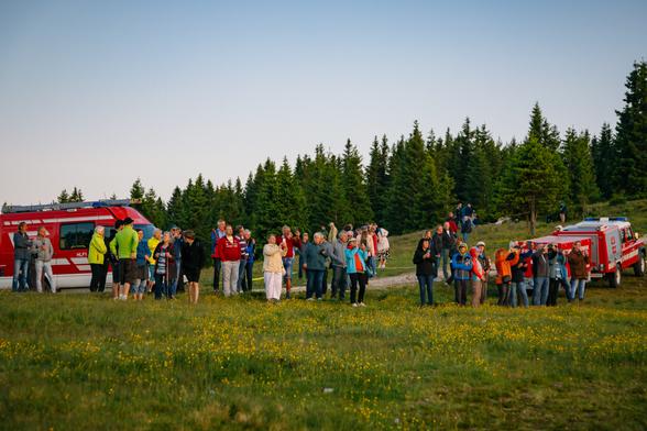 Dieses Bild zeigt eine größere Gruppe von Menschen, die sich auf einer Wiese in einer bergigen oder waldreichen Umgebung versammelt haben. Im Vordergrund sieht man eine Vielzahl von Personen unterschiedlichen Alters, viele tragen Outdoor- oder Wanderkleidung. Einige halten Kameras oder Smartphones in der Hand und machen Fotos vom Sonnenuntergang.
Im Hintergrund erkennt man mehrere rote Fahrzeuge der Feuerwehr, was auf eine organisierte Veranstaltung oder auf Sicherheitsbegleitung hindeuten könnte. Die Fahrzeuge stehen am Rand einer Wiese vor einem dichten Wald aus Nadelbäumen.
Die Atmosphäre wirkt ruhig und organisiert – die Leute stehen oder gehen entspannt umher, es scheint sich um eine friedliche Versammlung oder ein gemeinschaftliches Naturerlebnis zu handeln, etwa eine Sonnenaufgangswanderung oder eine Veranstaltung mit Feuerwehrpräsenz in den Bergen. Die Wiese ist mit kleinen gelben Blumen übersät, was dem Bild eine freundliche, sommerliche Stimmung verleiht.